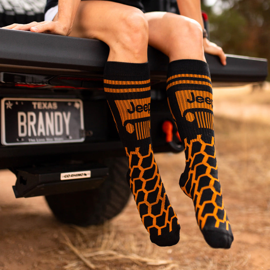 the lower half of a woman sitting on the back of a jeep wearing orange and black tread patterned socks with the jeep logo on them